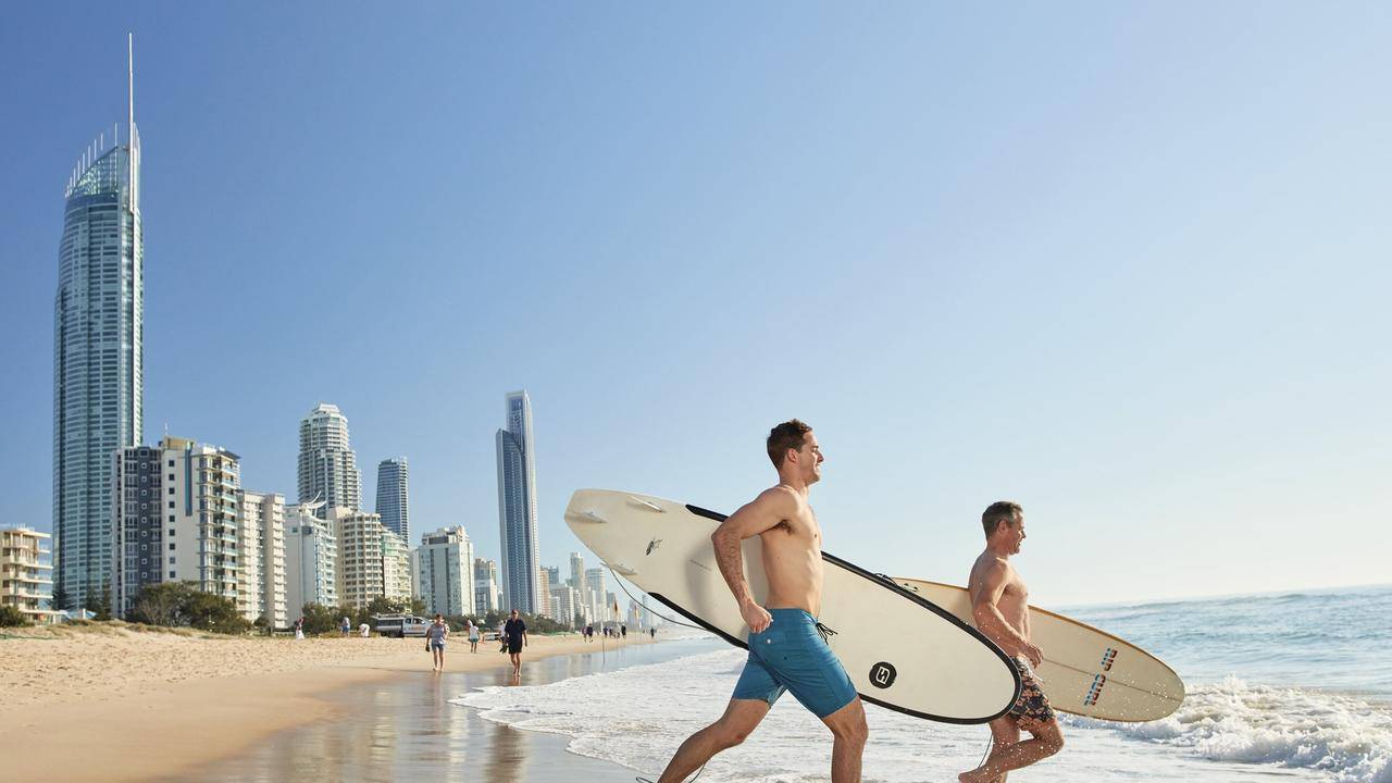 Two men carrying surfboards walking along the beach in the Gold Coast, with high-rise buildings and a clear blue sky in the background, highlighting the area's vibrant tourism and outdoor lifestyle.
